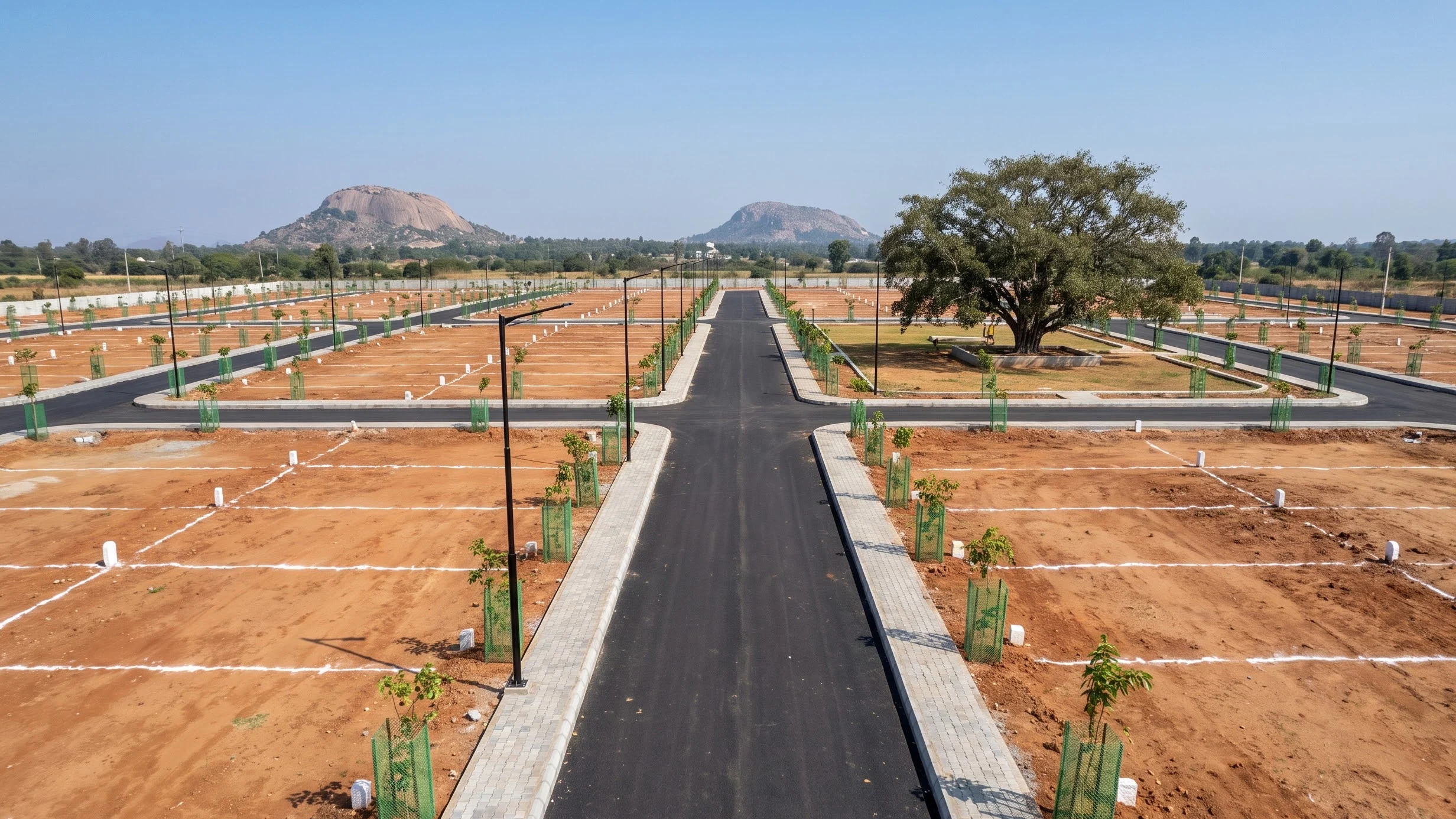 A plotted land development seen from above: a new road with footpaths, marked plots on red earth, young avenue trees and granite hills beyond