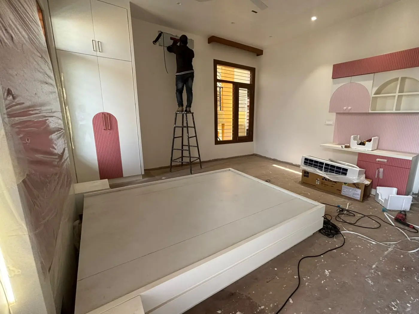 A child’s bedroom being fitted: a white wardrobe with a fluted pink arched panel, a pink and white study unit with arched shelves, a white bed base, and a fitter on a ladder at the far wall