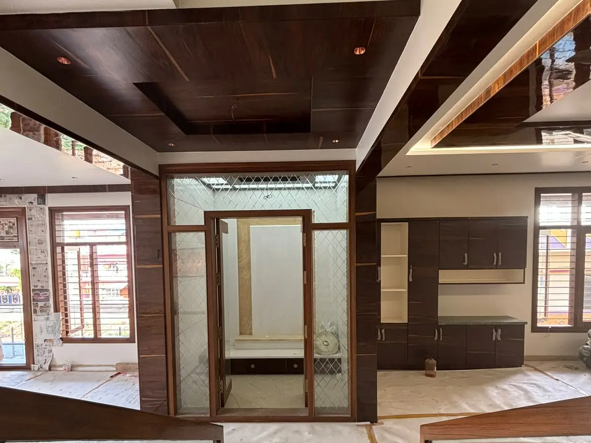 A glass-fronted puja room at the centre of a living floor, under a dark wood-finish ceiling, with a storage wall to its right; the floor is still covered in protective sheets