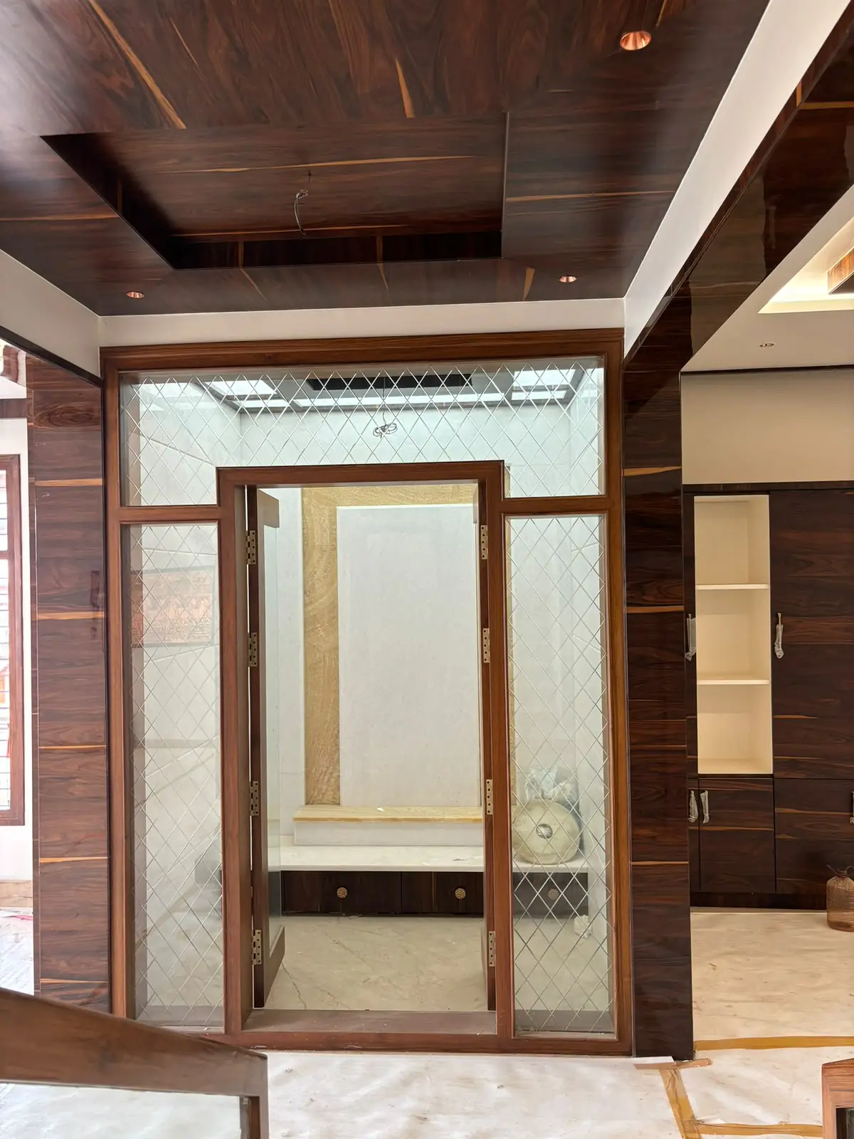 Puja room behind a timber frame glazed with diamond-etched glass: a stone-finish back panel, a platform with drawers below, and a coffered wood-finish ceiling outside it