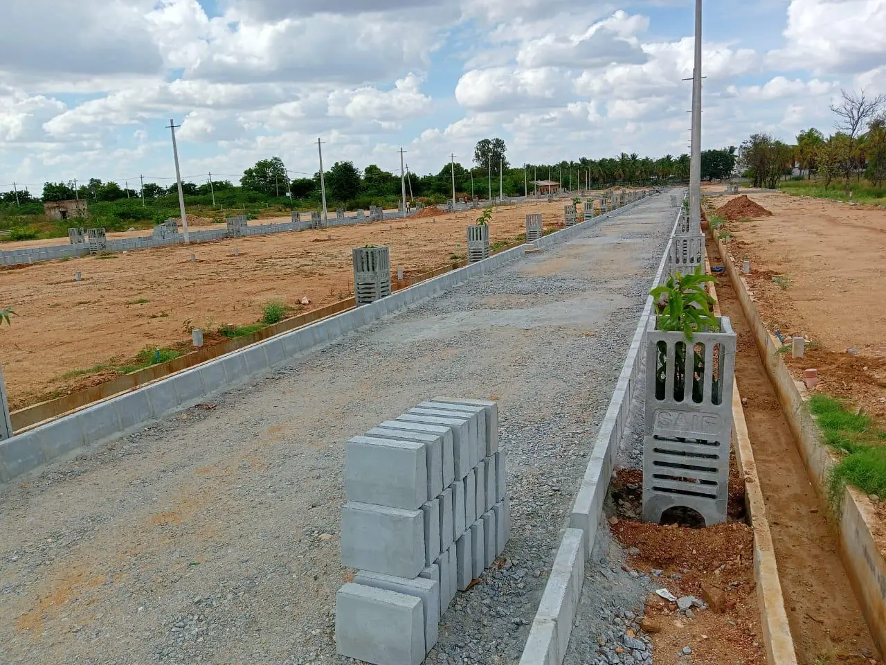 A stone road bed edged with kerbs, with precast drain units and a stack of kerb blocks waiting to be laid