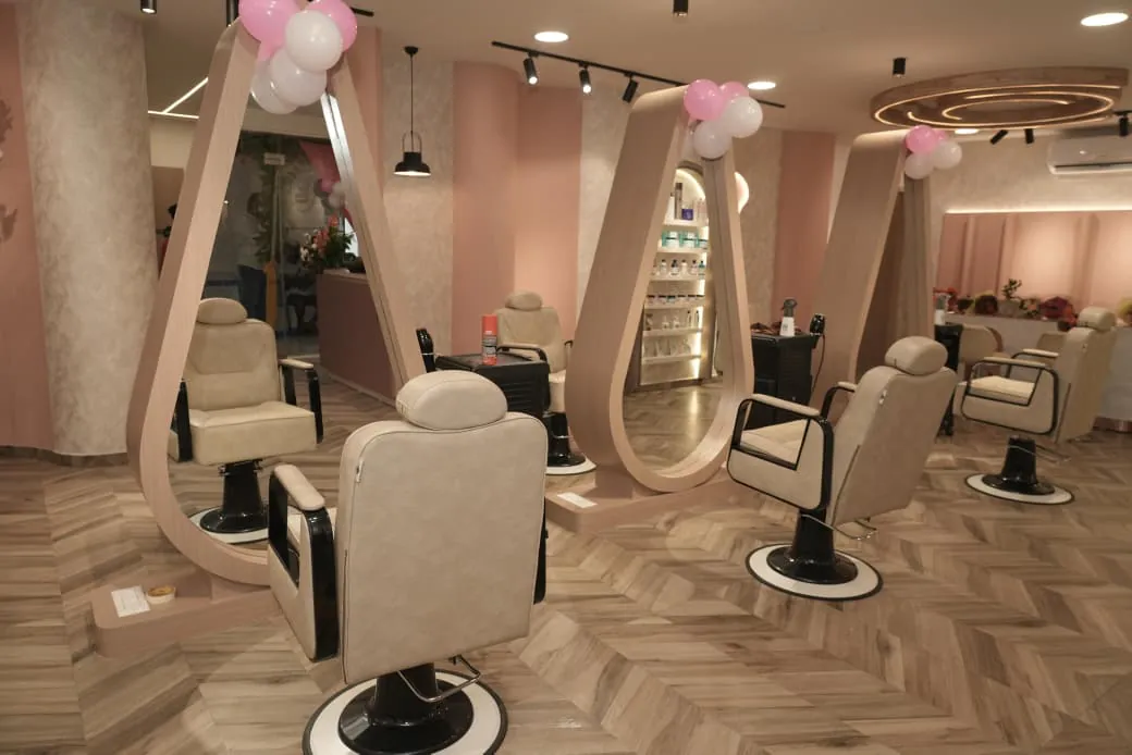 Salon styling floor: cream chairs facing tall teardrop mirrors in timber frames, on a herringbone floor with blush-pink walls and black track lights