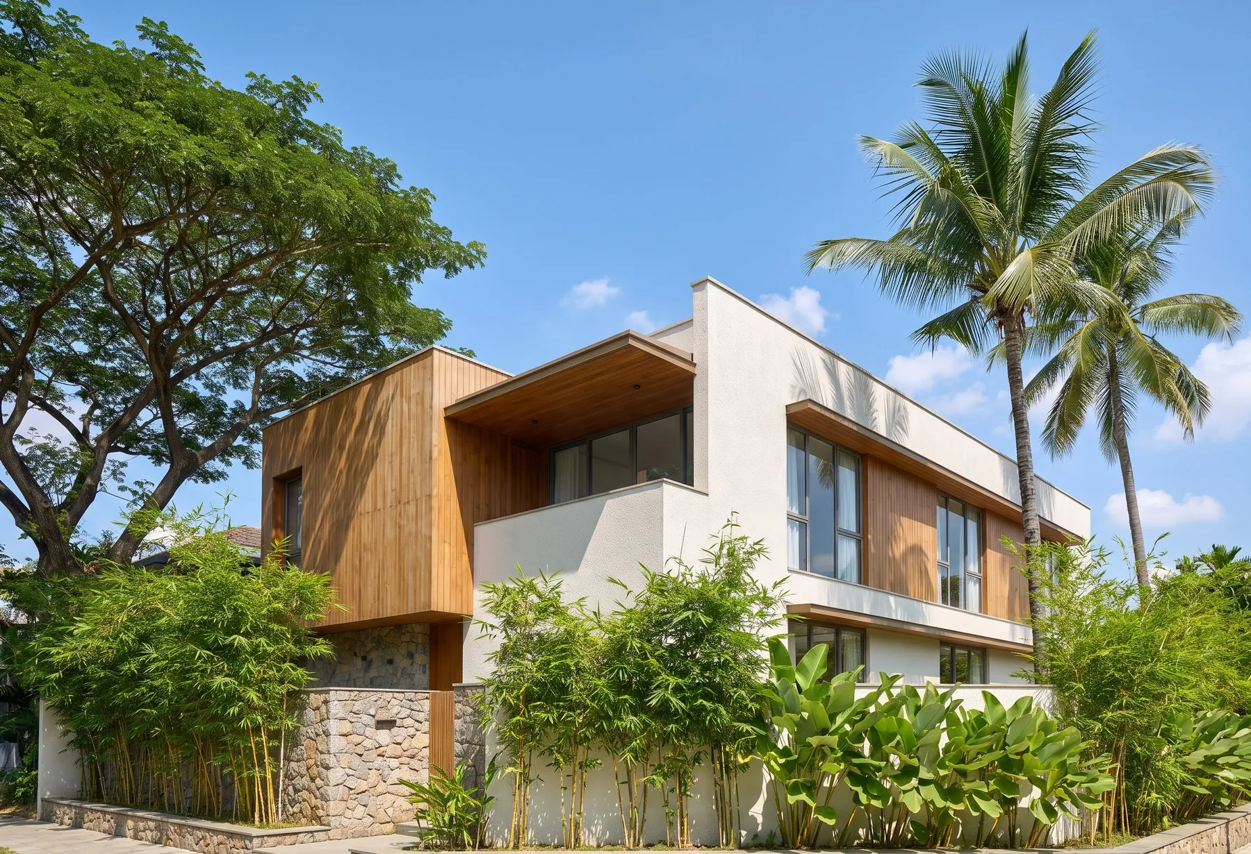 A finished two-storey home in timber, white plaster and stone behind a garden of bamboo, with a rain tree and coconut palms beside it