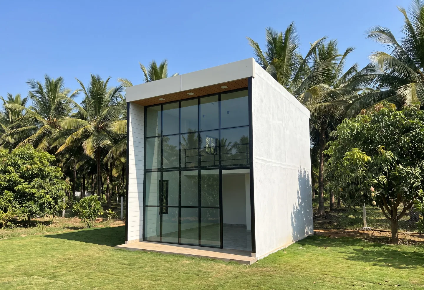 A tall box-shaped cabin with a full-height glazed front, on a lawn among mango trees and palms