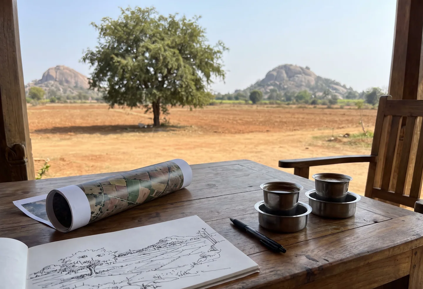 A table on a verandah with a rolled survey map and a sketchbook, looking out over open land to granite hills