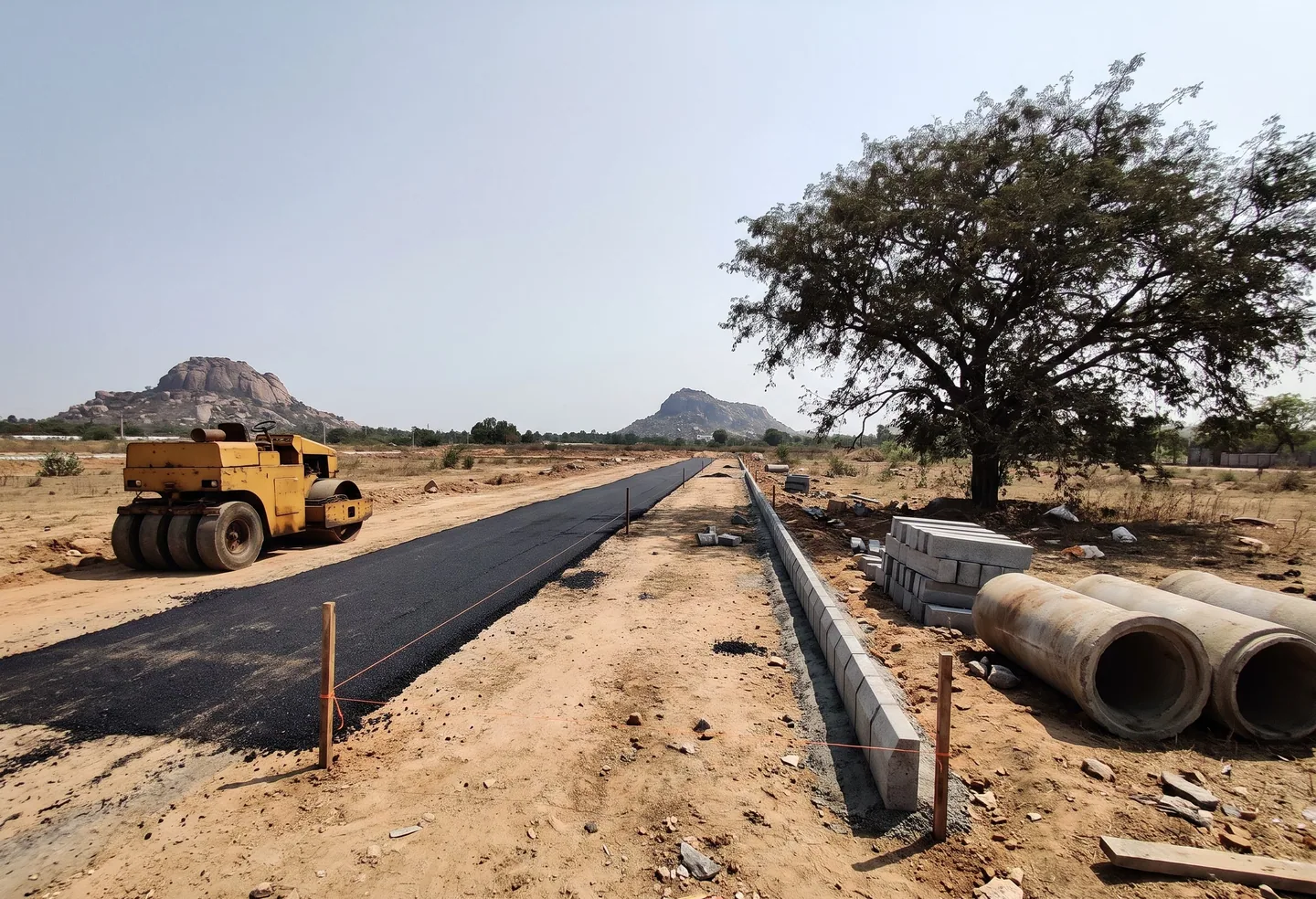 A new road being laid across open land: fresh asphalt, a road roller, kerb stones and drain pipes
